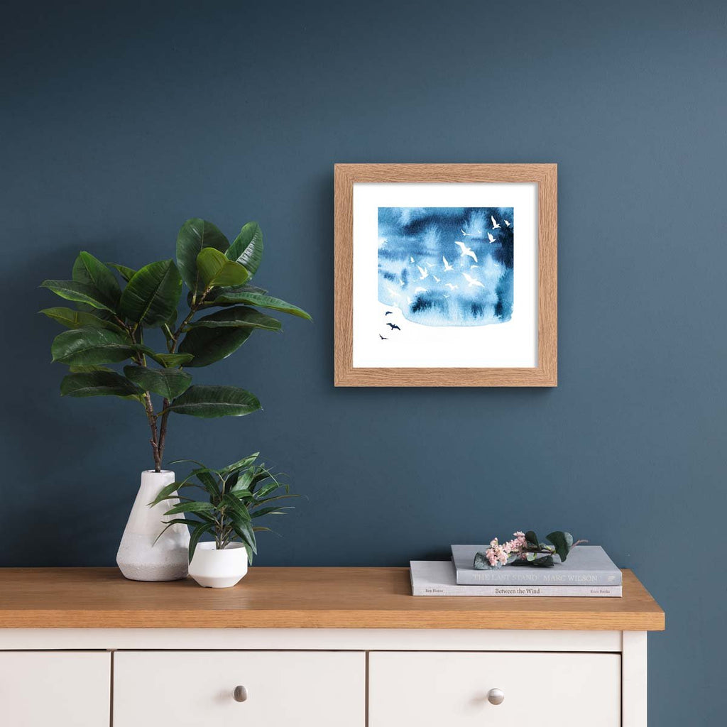 Watercolour art print featuring the silhouettes of birds flying against a brilliant blue sky. Art print is  hung on a dark blue wall.