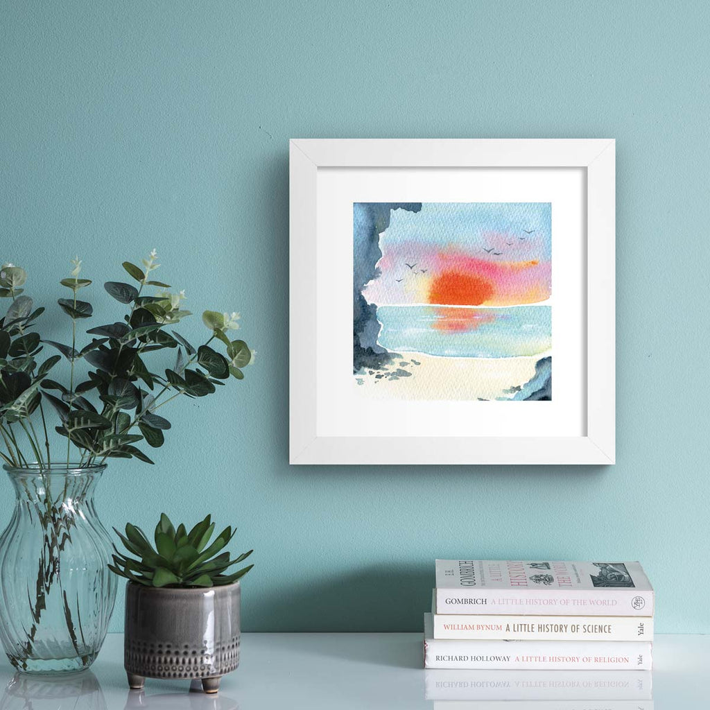 Vibrant watercolour art print featuring a sunset over a coastal scene. Art print is hung up on a pale blue wall.