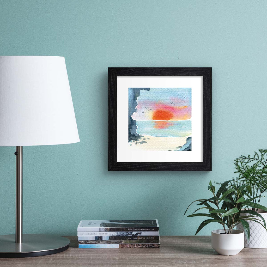 Vibrant watercolour art print featuring a sunset over a coastal scene. Art print is hung up on a blue wall.