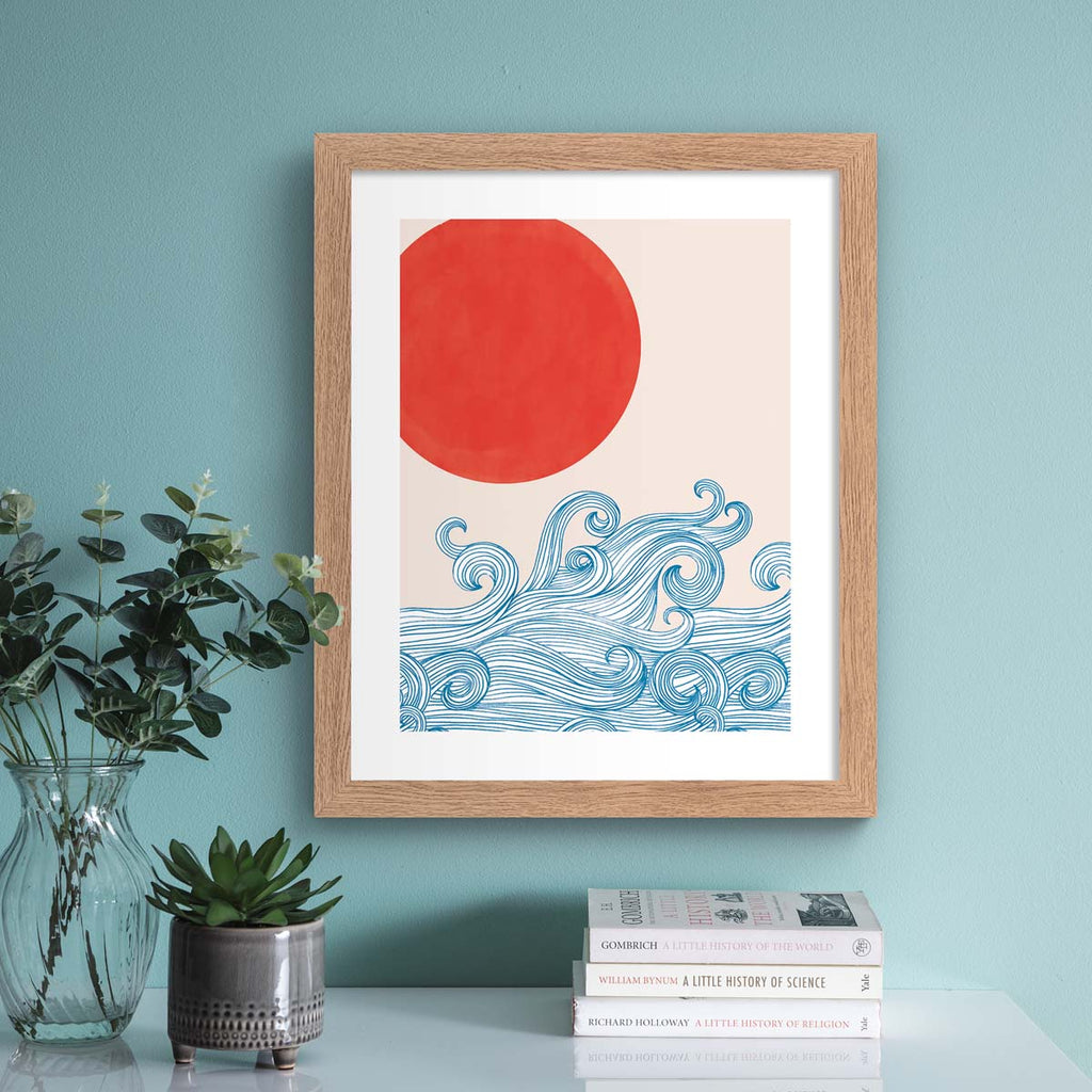 Beautiful art print featuring waves reaching up to a red sun. Art print is hung up on a light blue wall.