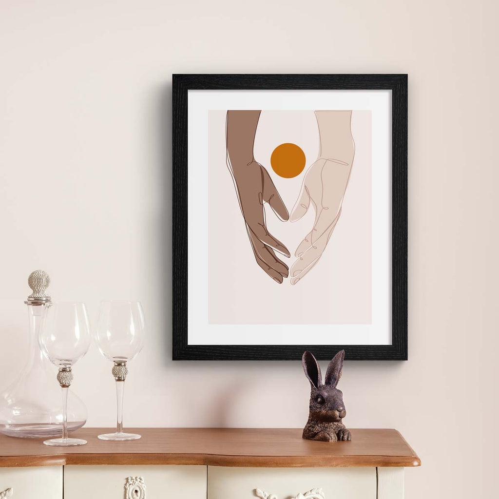 Beautiful art print featuring two hands meeting in the meeting of the frame. A bright sun sits behind the hands. Art print is hung on a beige wall.