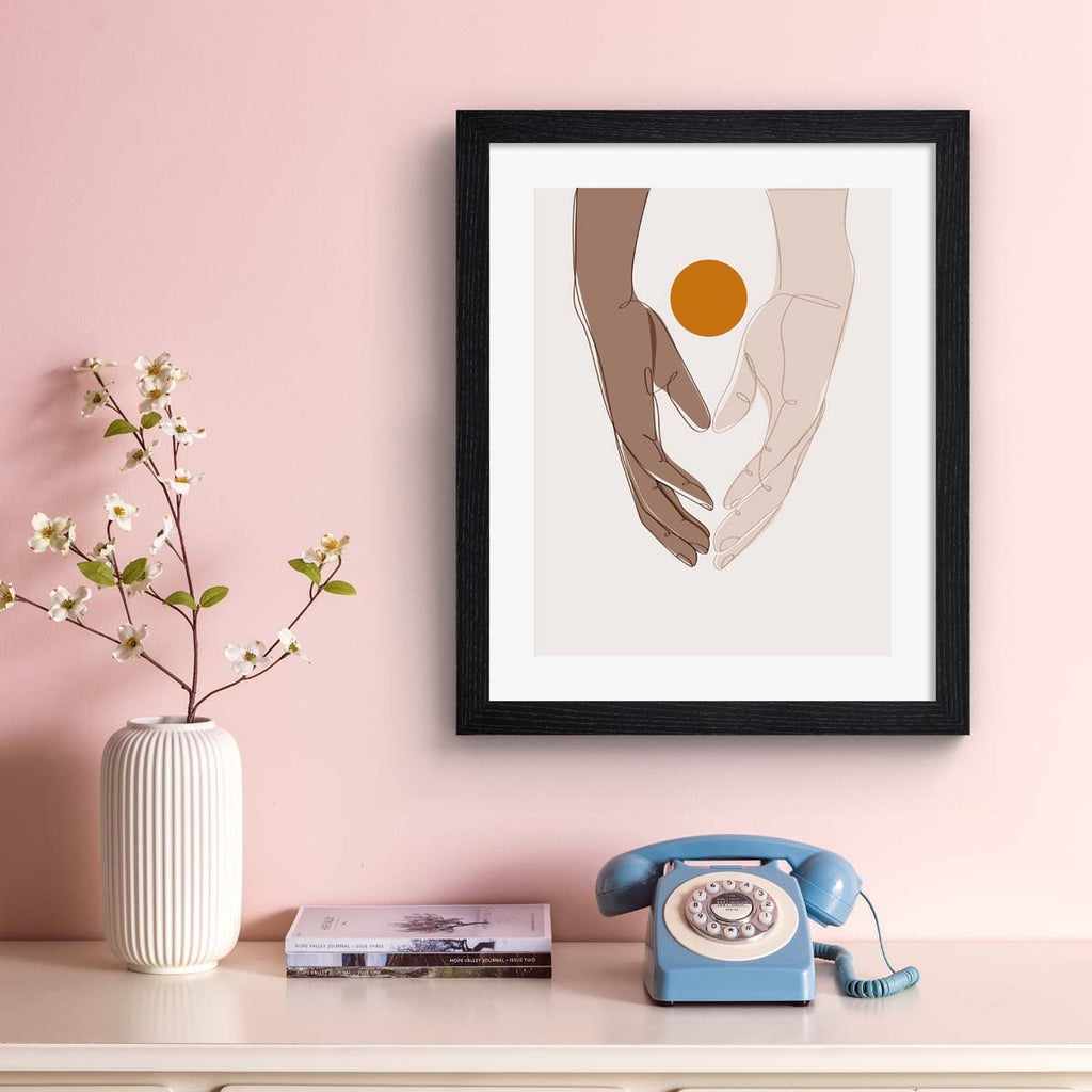 Beautiful art print featuring two hands meeting in the meeting of the frame. A bright sun sits behind the hands. Art print is hung on a pale pink wall.