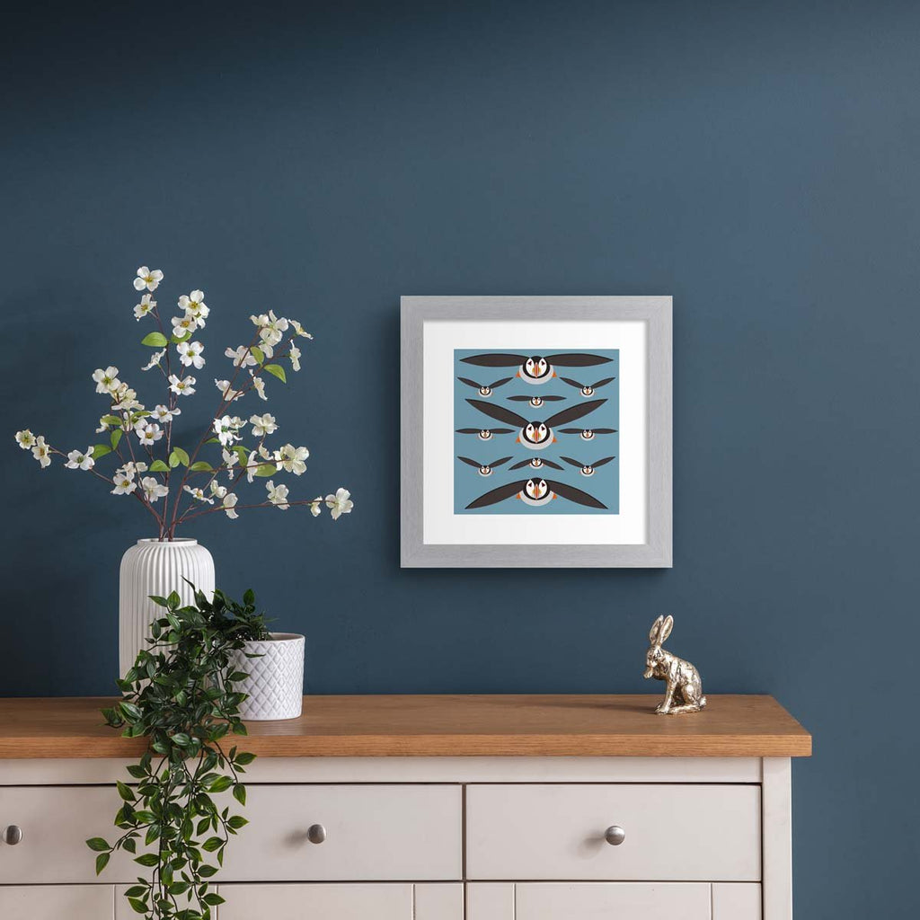 This minimalistic art print features a brilliant pattern of puffins flying in formation. Art print is hung up on a dark blue wall.