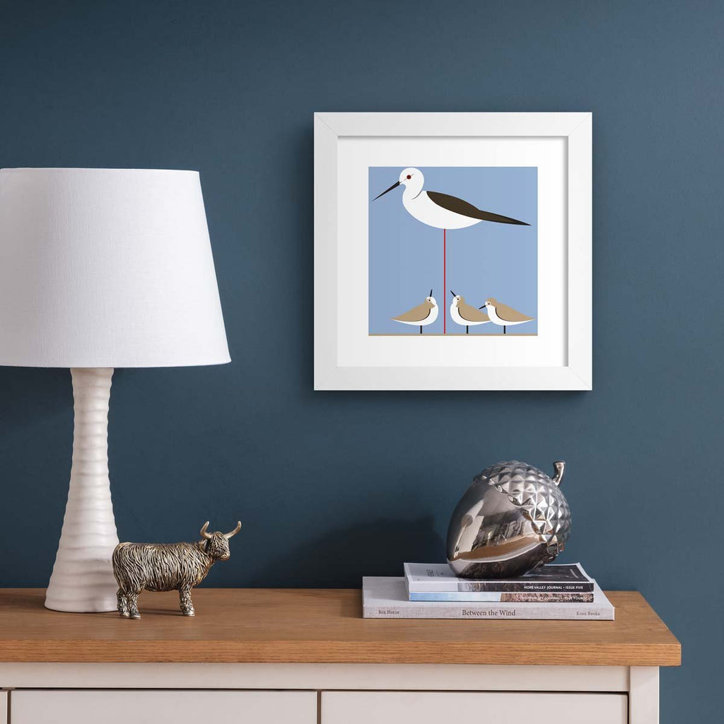 Minimalistic art print featuring small birds perched underneath a tall stilt. Art protein is hung up on a dark blue wall.