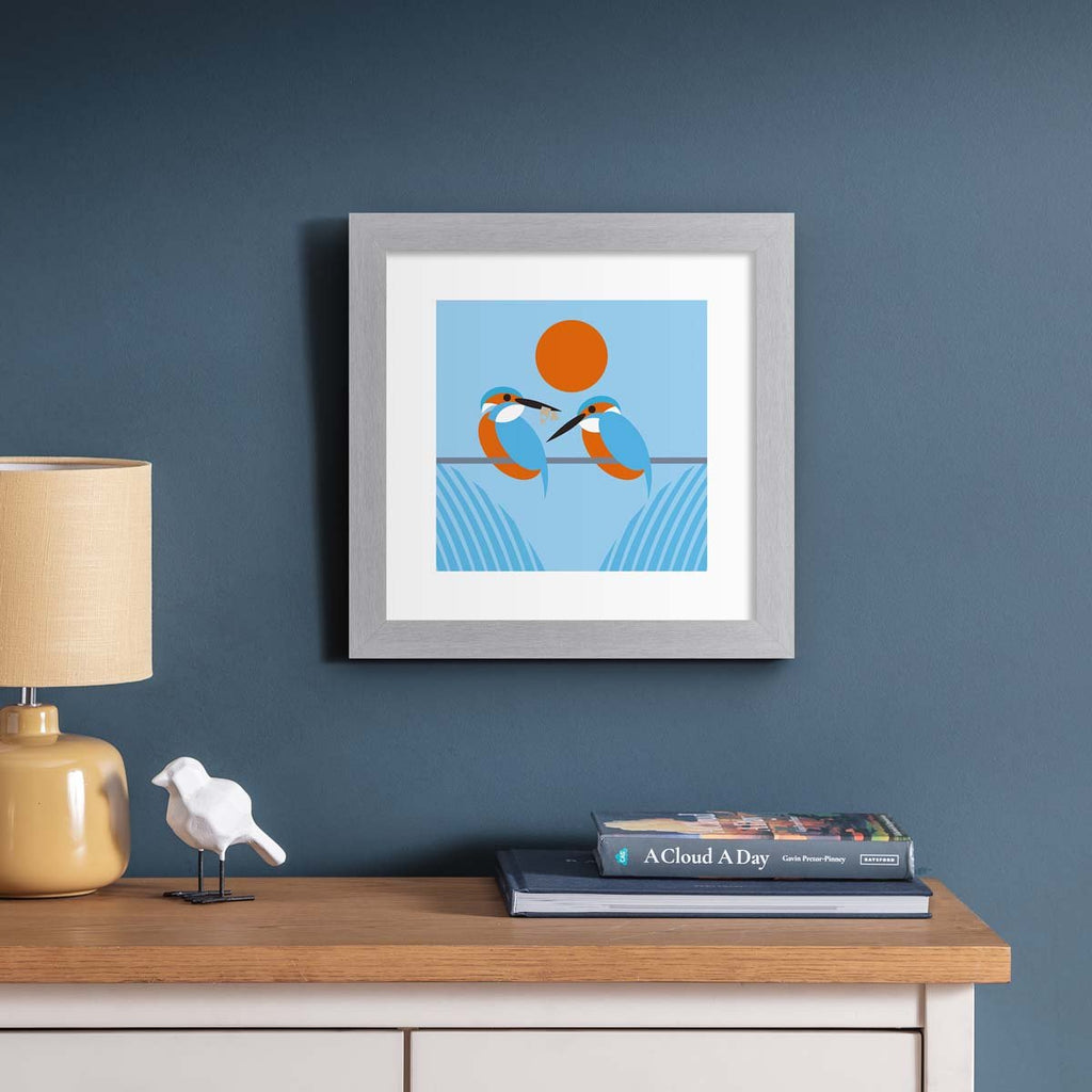 Minimalistic art print featuring two beautiful kingfishers perched on a line, framed by a red sunset. Art print is hung up on a dark blue wall.
