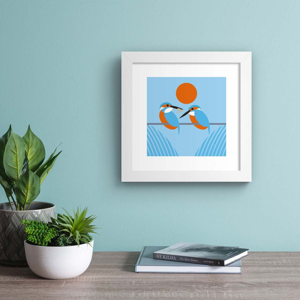 Minimalistic art print featuring two beautiful kingfishers perched on a line, framed by a red sunset. Art print is hung up on a blue wall.