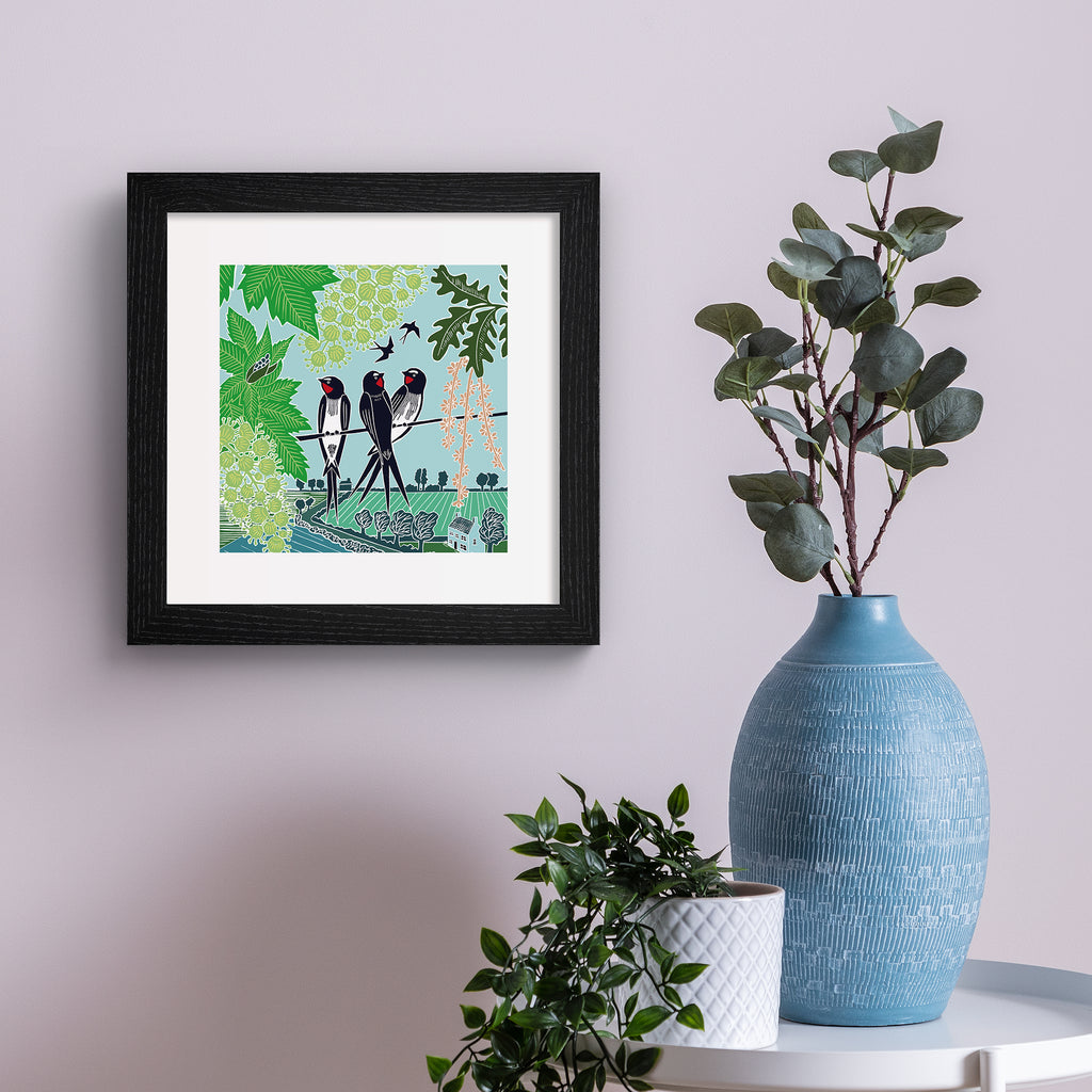 Emotive art print featuring three swallows perching on a branch above a beautiful Summer nature scene. Art print is hung up on a pink wall.