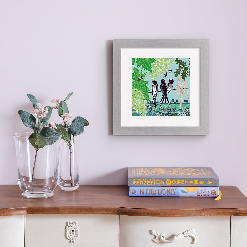 Emotive art print featuring three swallows perching on a branch above a beautiful Summer nature scene. Art print is hung up on a pale pink wall.