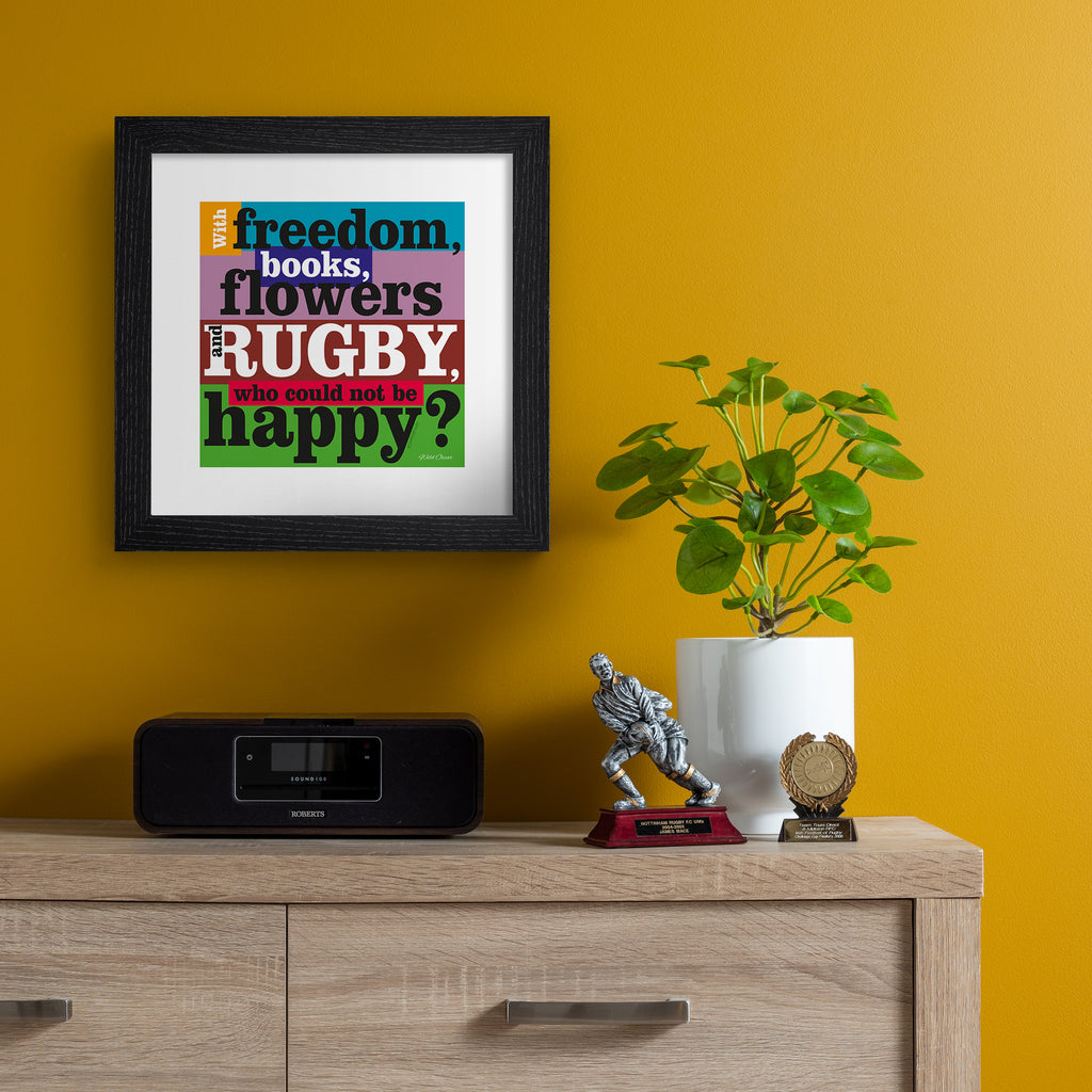 Art print featuring typography celebrating rugby, on a bright multicoloured background. Typography reads 'With Freedom, Books, Flowers And Rugby, Who Could Not By Happy'. Art print is hung up on an orange wall.