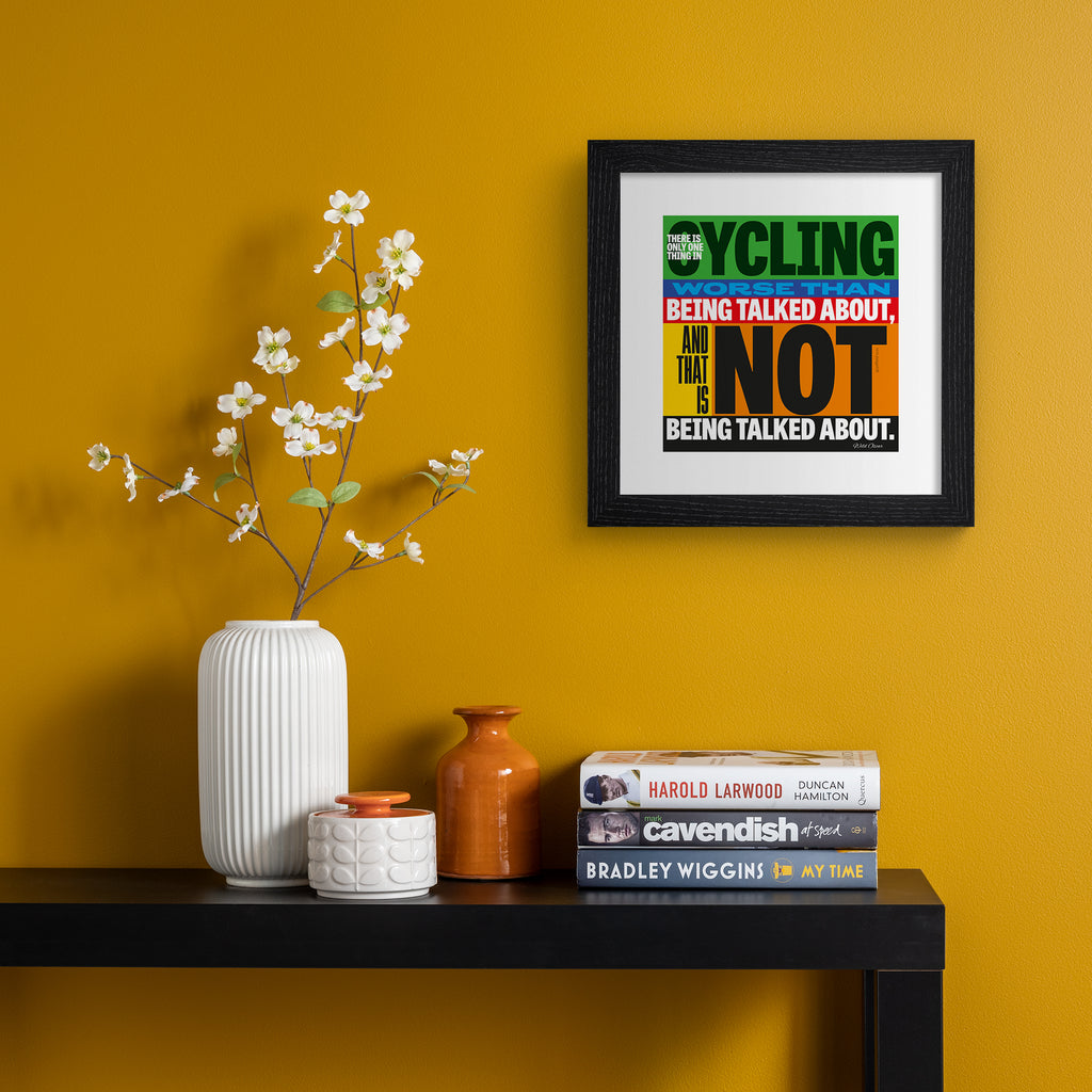 Sporty art print featuring typography celebrating cycling, on a multicolour background. Typography reads 'There Is Only One Thing In Cycling Worse Than Being Talked About, And That Is Not Being Talked About.' Art print is hung up on an orange wall.