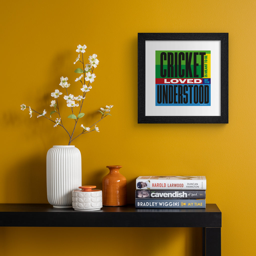Typography art print celebrating cricket, on a bright green, blue, yellow and red background. Typography reads 'Cricket Is Meant To be Loved, Not To Be Understood'. Art print is hung up on an orange wall.