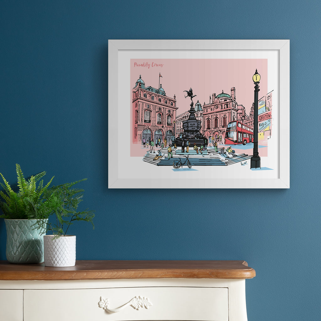 Vibrant travel art print featuring a detailed illustration of Picadilly Circus in London, in front of a pale pink background. Title in top left hand corner reads 'Piccadilly Circus'. Art print is hung up on a dark blue wall.