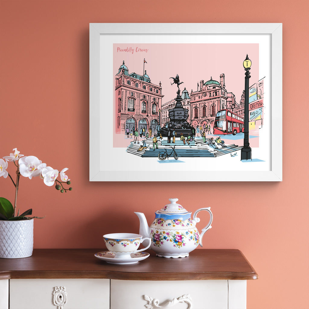 Vibrant travel art print featuring a detailed illustration of Picadilly Circus in London, in front of a pale pink background. Title in top left hand corner reads 'Piccadilly Circus'. Art print is hung up on a pink wall.