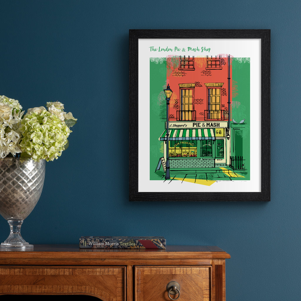 Vibrant travel art print featuring the London Pie & Mash Shop, amidst a bright orange and green background. Title at the top reads 'The London Pie & Mash Shop'. Art print is hung up on a dark blue wall.