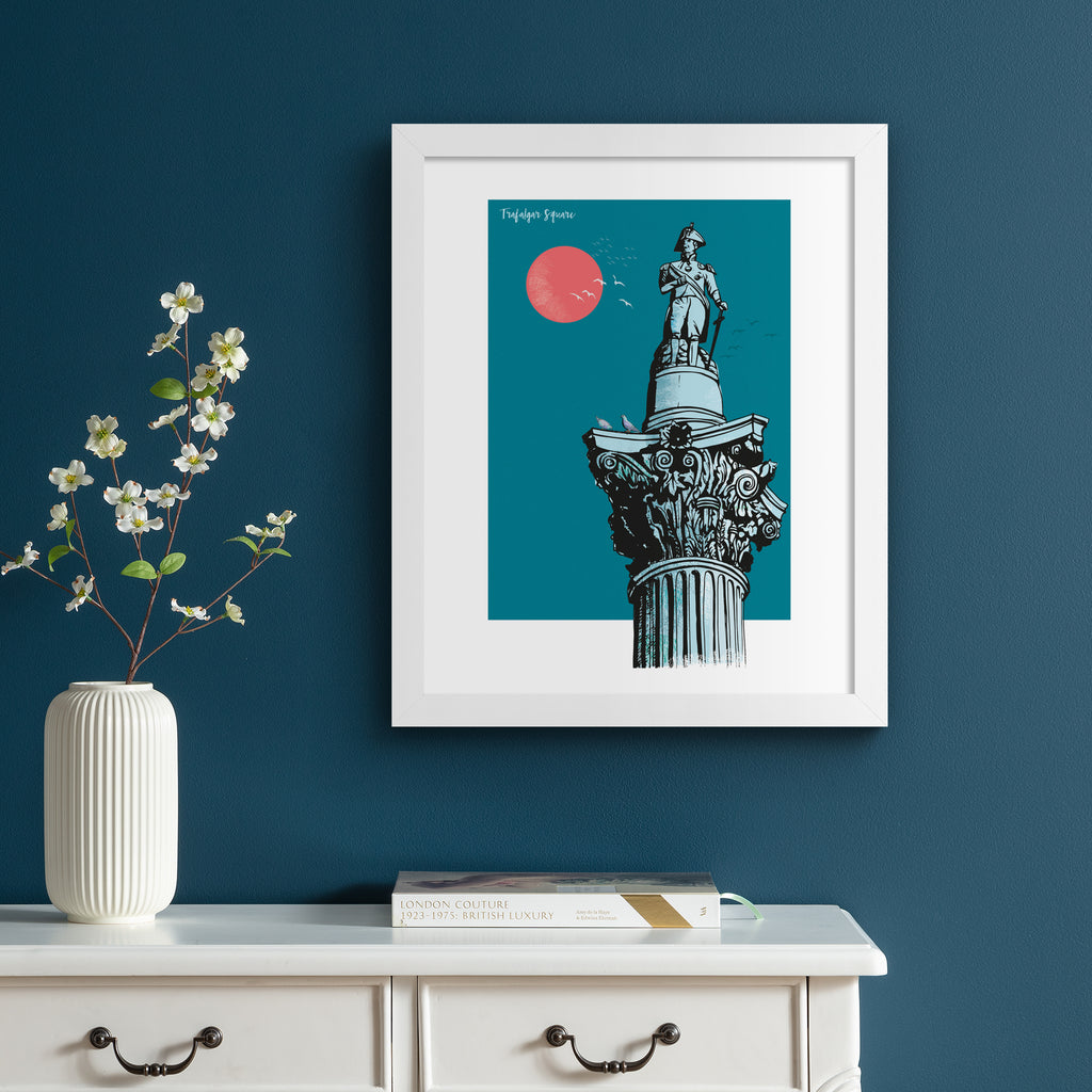 Vibrant travel art print featuring Trafalgar Square in London, in front of a brilliant blue sky and red sun. Title in the top left reads 'Trafalgar Square'.  Art print is hung up on a dark blue wall.