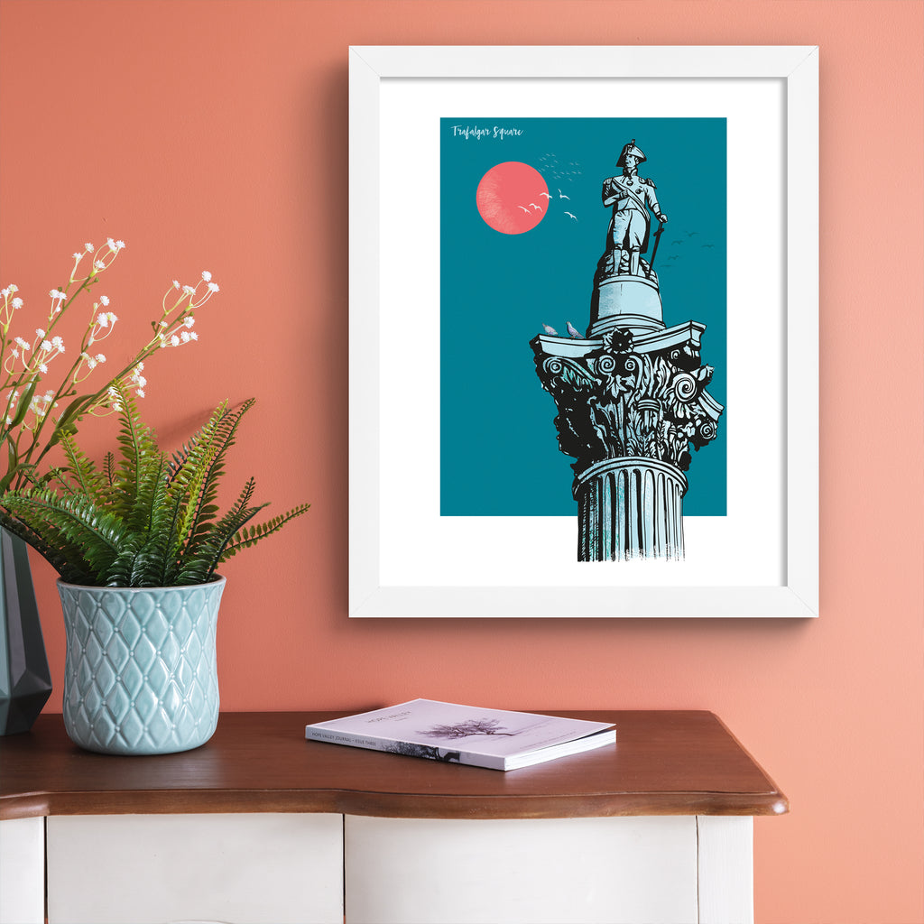 Vibrant travel art print featuring Trafalgar Square in London, in front of a brilliant blue sky and red sun. Title in the top left reads 'Trafalgar Square'.  Art print is hung up on a pink wall.