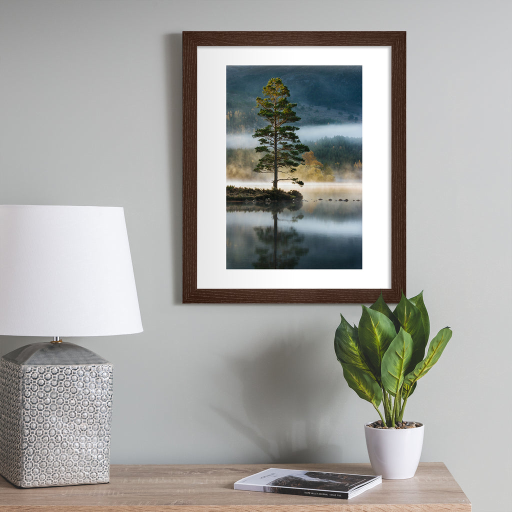 Photography art print featuring a tree standing on the edge of a reflective lake, bordered by a misty forest. Art print is hung up on a grey wall.