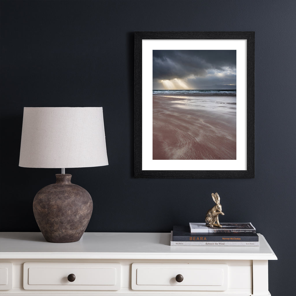 Photography art print featuring a coastal view of the the beach and the sea, with beams of light peeking through moody clouds. Art print is hung up on a dark blue wall.