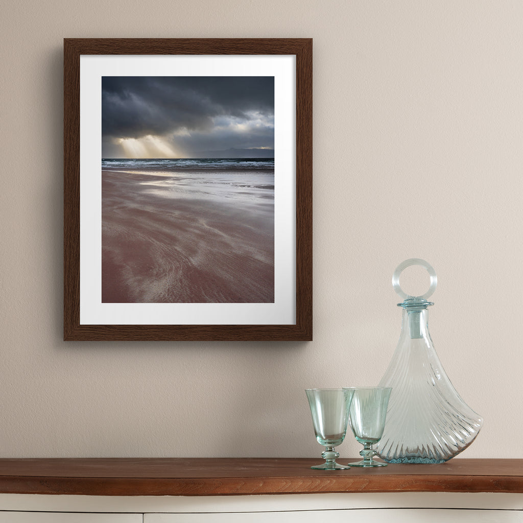 Photography art print featuring a coastal view of the the beach and the sea, with beams of light peeking through moody clouds. Art print is hung up on a beige wall.