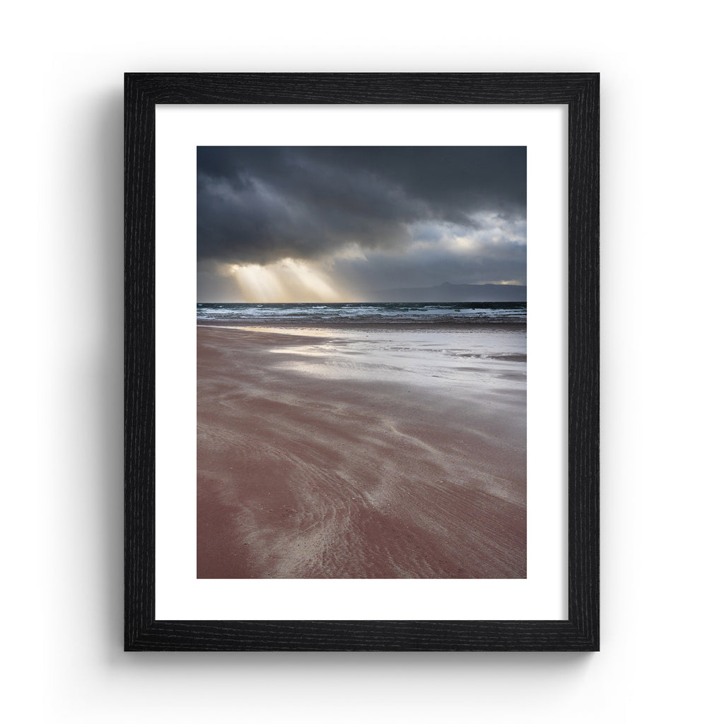 Photography art print featuring a coastal view of the the beach and the sea, with beams of light peeking through moody clouds. Art print is in a black frame.