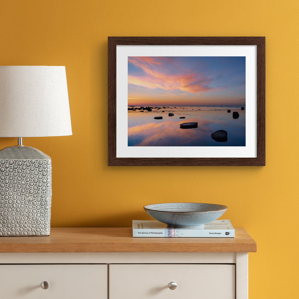 Stunning photography art print featuring a vivid sunrise over a reflective beach. Art print is hung up on an orange wall.