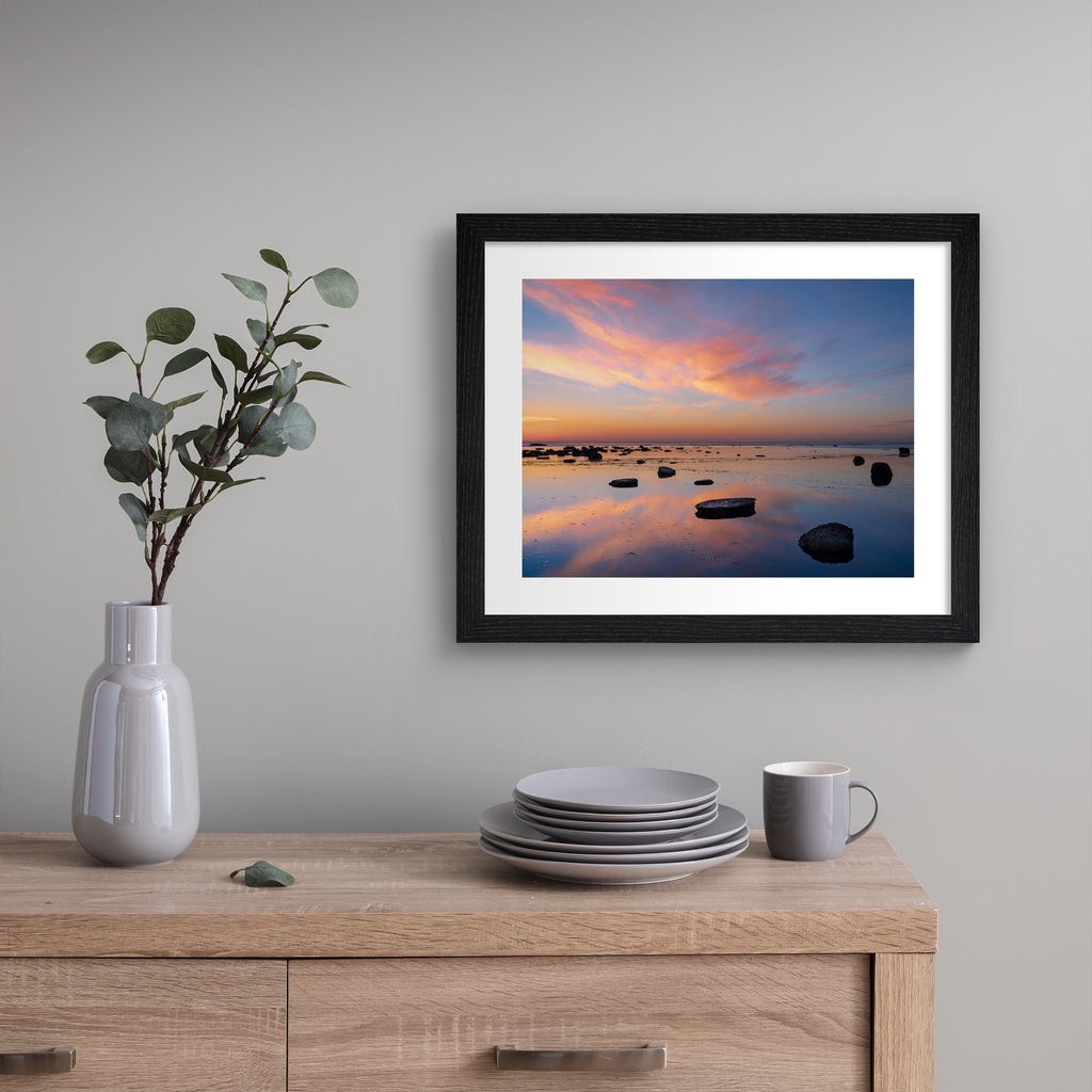 Stunning photography art print featuring a vivid sunrise over a reflective beach. Art print is hung up on a grey wall.
