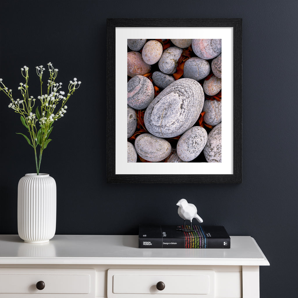 Beautifully simplistic photography art print featuring a close of up of pebbles found on a beach. Art print is hung up on a dark blue wall.