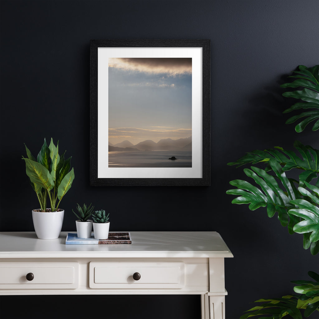 Emotive photography art print featuring a sweeping scene of the Isle of Sky, including shimmering water, a warm sunrise and moody cliffs in the distance. Art print is hung up on a navy blue wall.