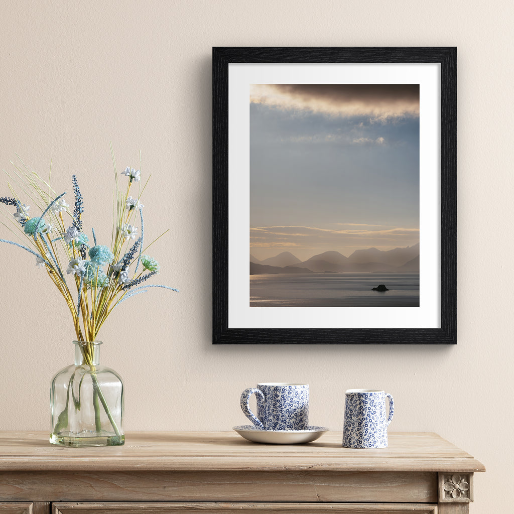 Emotive photography art print featuring a sweeping scene of the Isle of Sky, including shimmering water, a warm sunrise and moody cliffs in the distance. Art print is hung up an a beige wall.