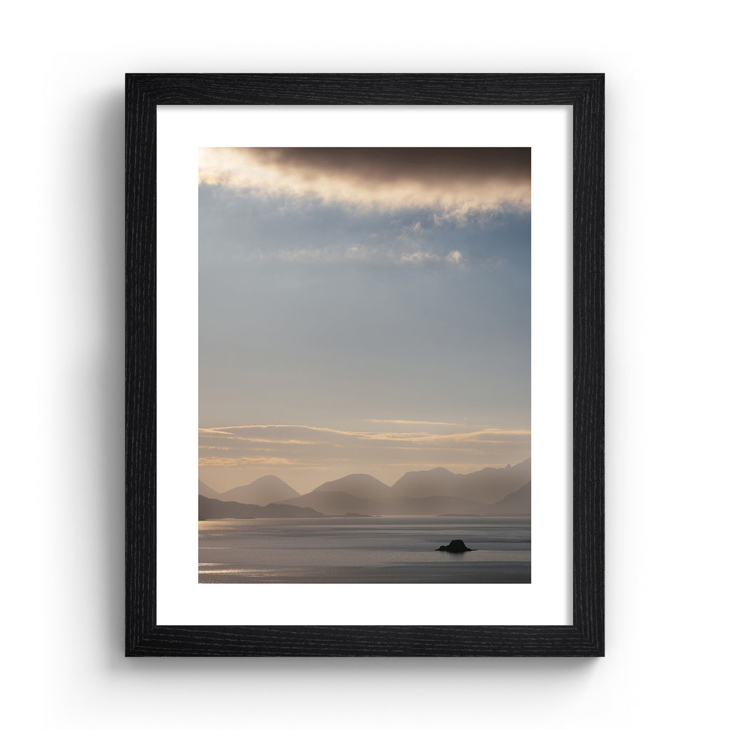 Emotive photography art print featuring a sweeping scene of the Isle of Sky, including shimmering water, a warm sunrise and moody cliffs in the distance. Art print is in a black frame.