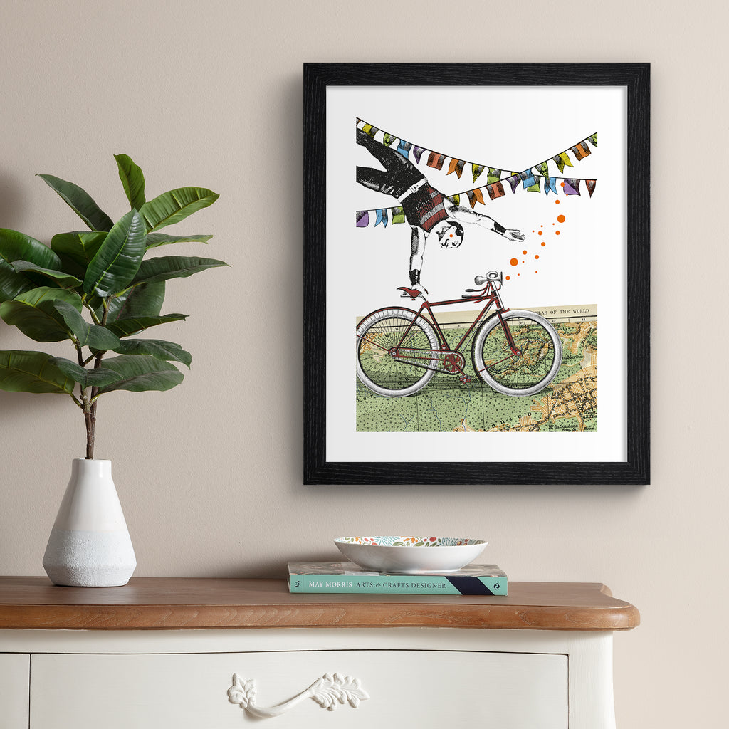 Eclectic art print featuring a man balancing off of a bicycle, perched on a map of the world. Art print is hung up on a beige wall.