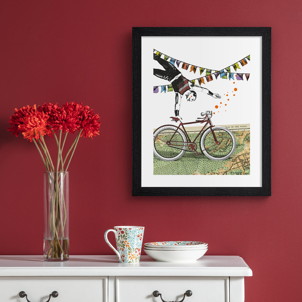 Eclectic art print featuring a man balancing off of a bicycle, perched on a map of the world. Art print is hung up on a red wall.