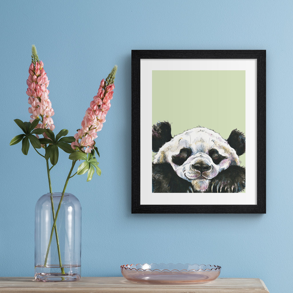 Playful animal art print featuring a happy panda popping into the frame, in front of a sage green background. Art print is hung up on a blue wall.