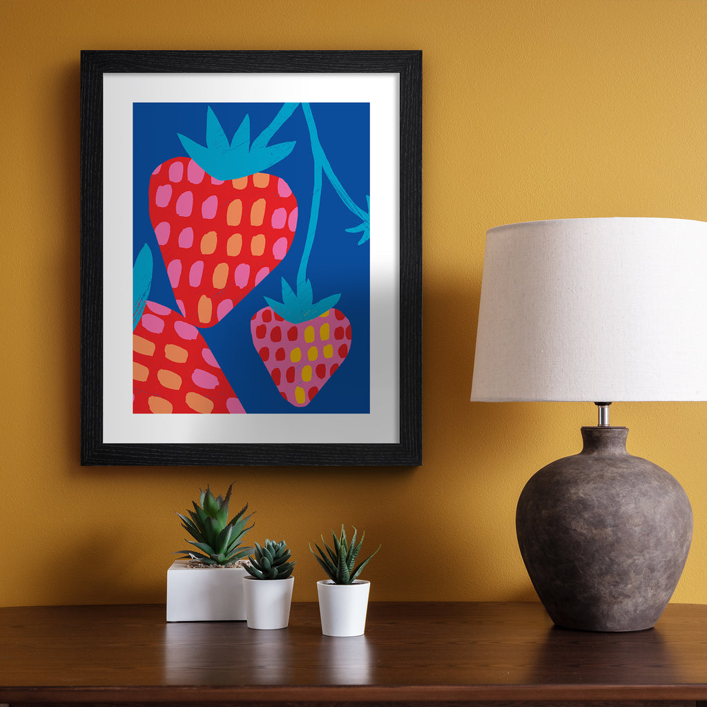 Vivid art print containing a bunch of strawberries dangling in front of a bright blue background. Art Print is hung up on an orange wall.