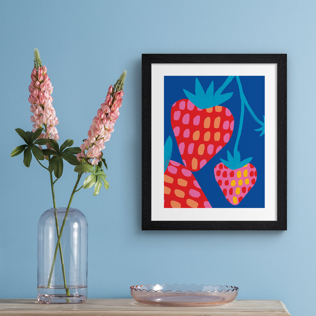 Vivid art print containing a bunch of strawberries dangling in front of a bright blue background. Art print is hung up on a blue wall.