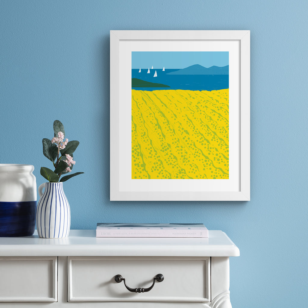 Minimalistic art print featuring beautiful yellow fields bordered by a coastal view.  Art print is hung up on a pale blue wall.