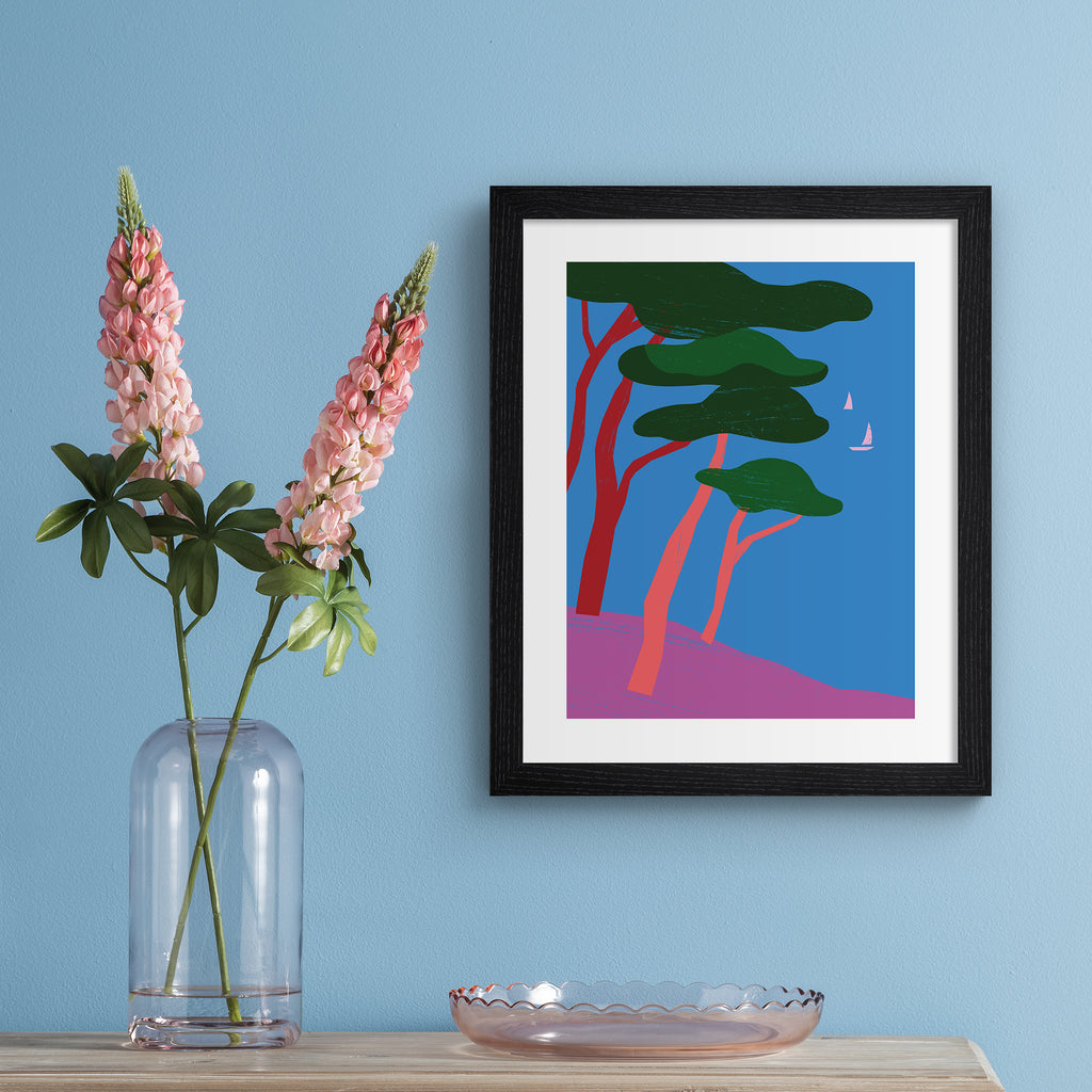 Bright art print featuring a beautiful tropical scene of trees overlooking the sea, with sailboats bobbing in the distance. Art print is hung up on a blue wall.