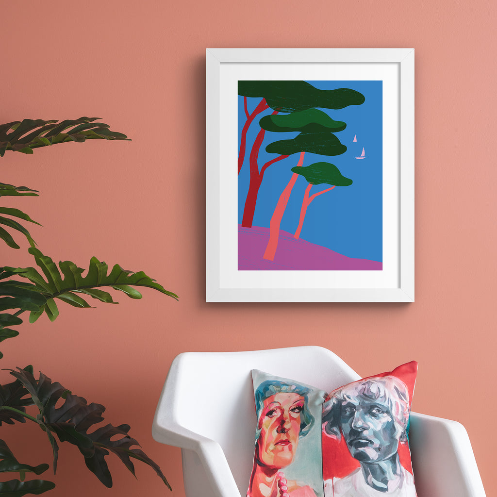 Bright art print featuring a beautiful tropical scene of trees overlooking the sea, with sailboats bobbing in the distance. Art print is hung up on a pink wall.