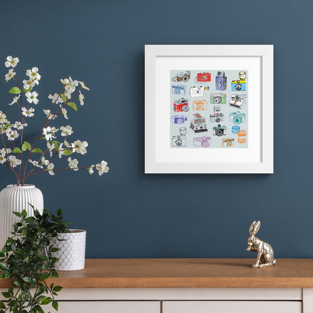 Playful art print featuring different types of retro cameras on a pale blue background. Art print is hung up on a dark blue wall.