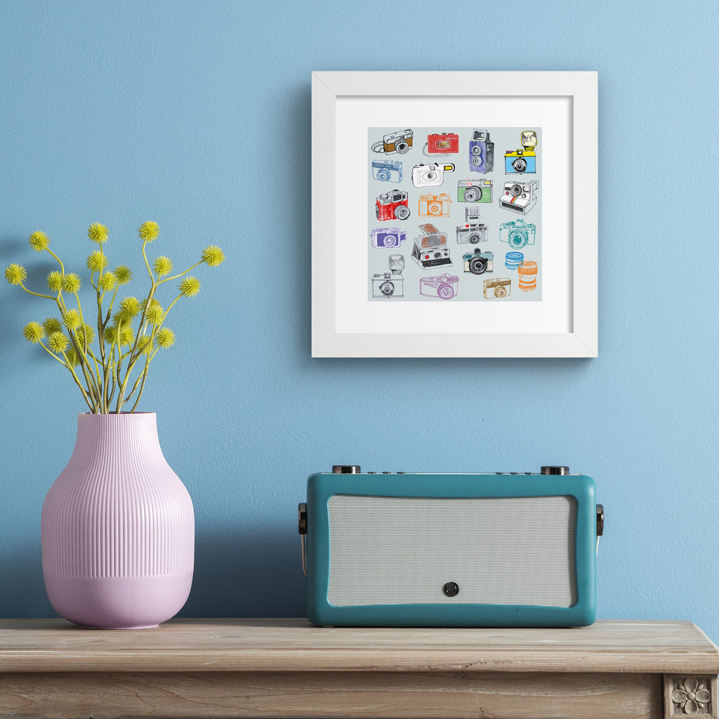 Playful art print featuring different types of retro cameras on a pale blue background. Art print is hung up on a pale blue wall.
