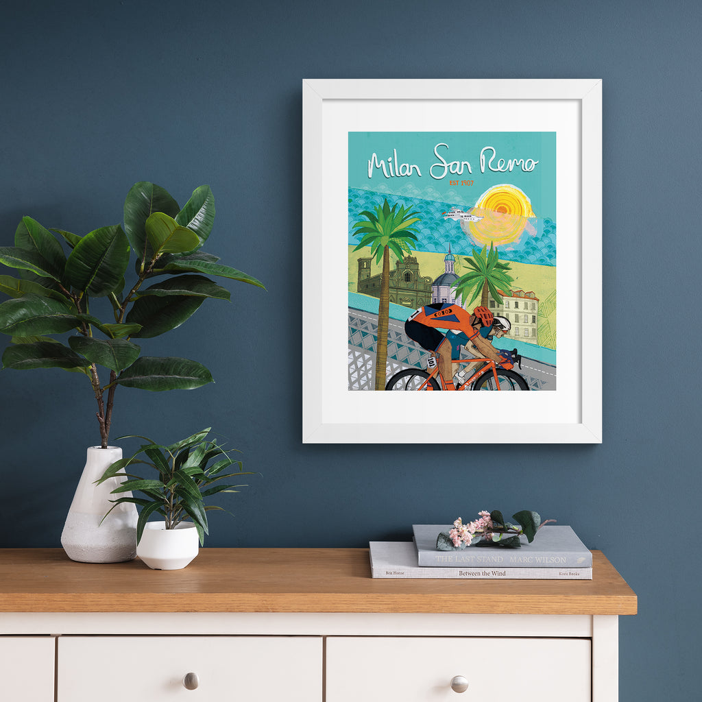 Colourful art print featuring cyclists in front of a beautiful coastal city. Text above reads 'Milan San Remo'. Art print is hung up on a dark blue wall.