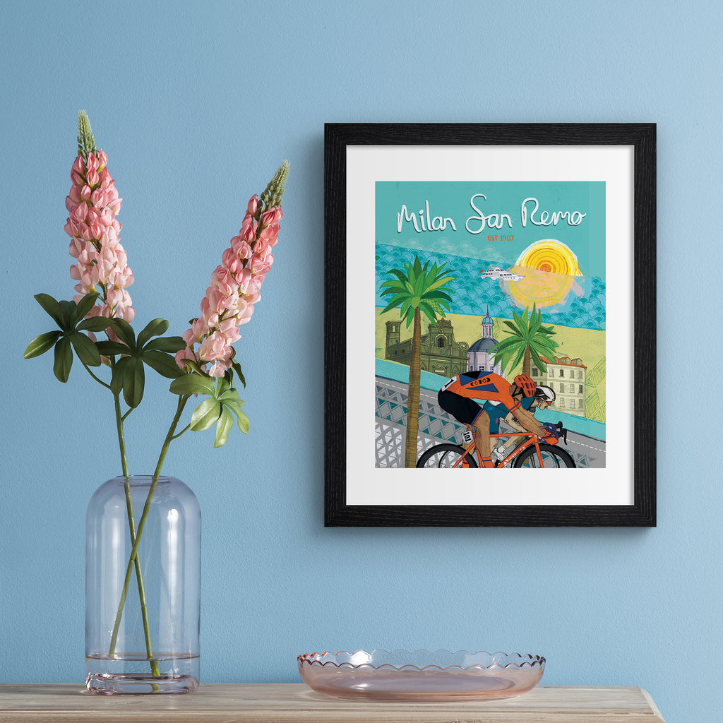 Colourful art print featuring cyclists in front of a beautiful coastal city. Text above reads 'Milan San Remo'. Art print is hung up on a light blue wall.