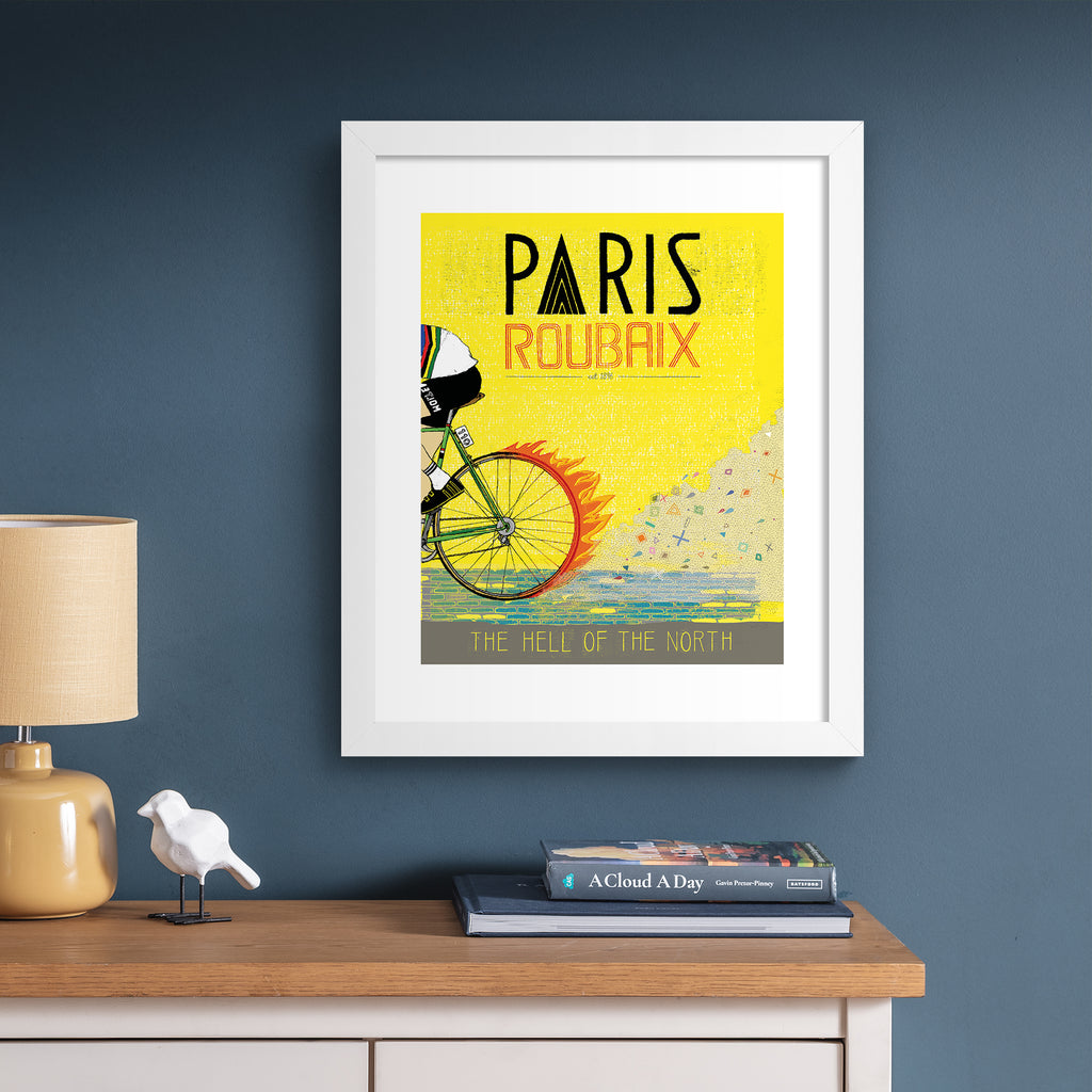 Bright art print featuring a person cycling past on a yellow background. Title above reads 'Paris, Roubaix'. Art print is hung up on a dark blue wall.