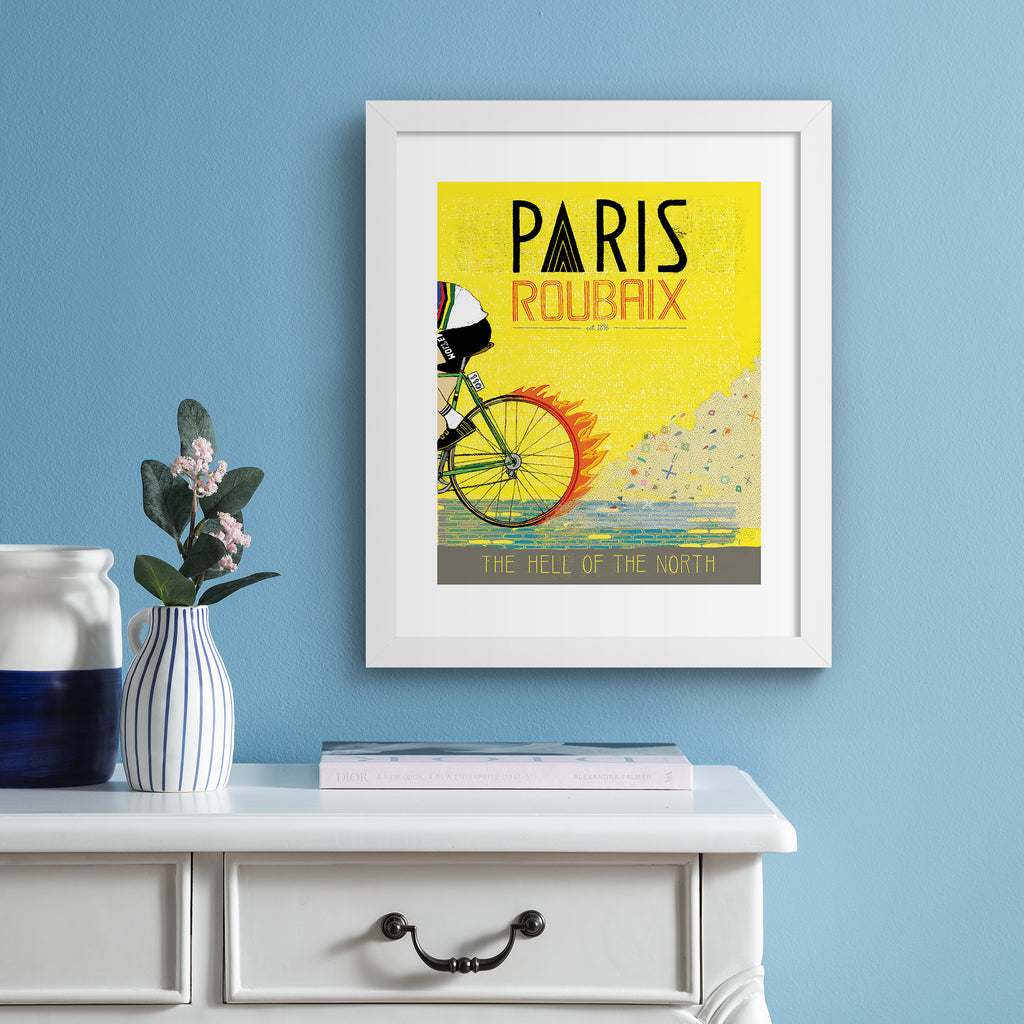 Bright art print featuring a person cycling past on a yellow background. Title above reads 'Paris, Roubaix'. Art print is hung up on a light blue wall.