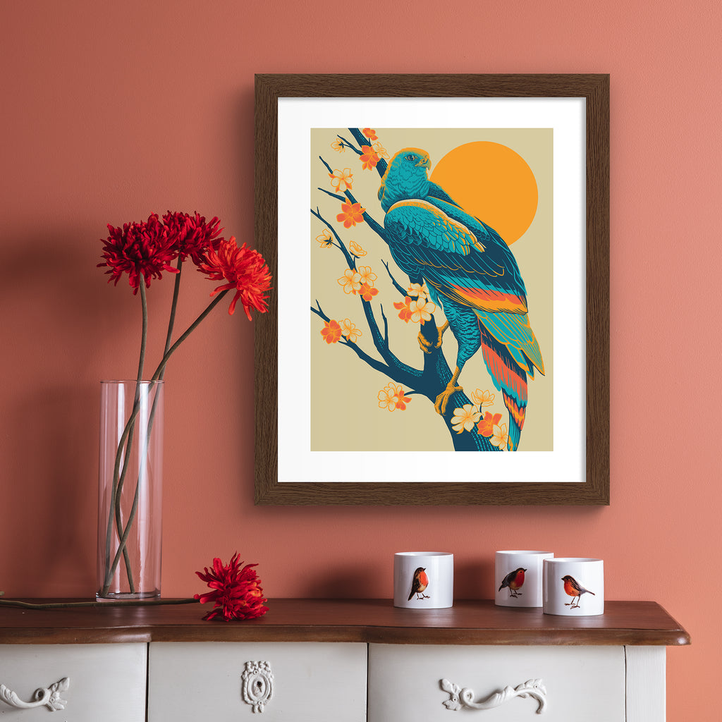Colourful print featuring a detailed illustration of a peregrine falcon perched in front of a brilliant sunrise. Art print is hung up on a pink wall.