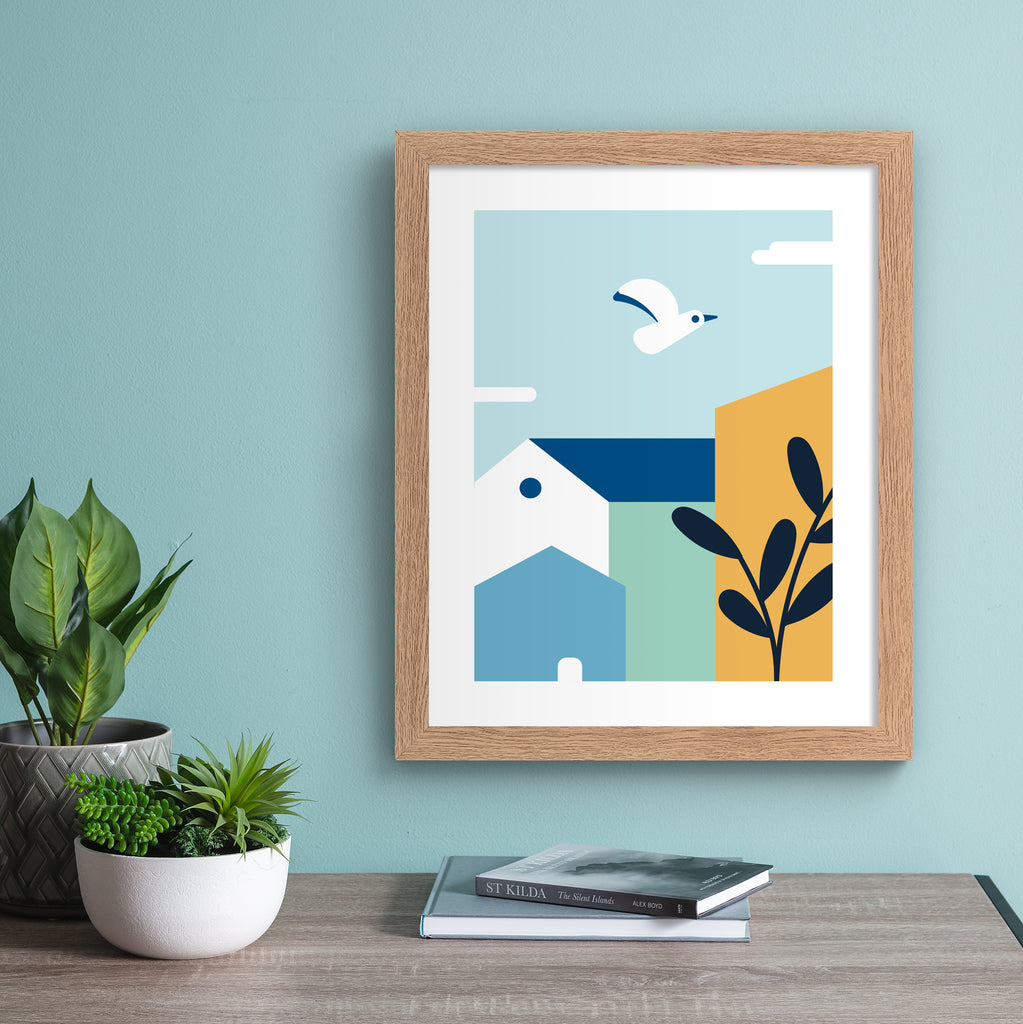 Minimalistic art print featuring a coastal bird flying over colourful houses. Art print is hung up on a light blue wall.