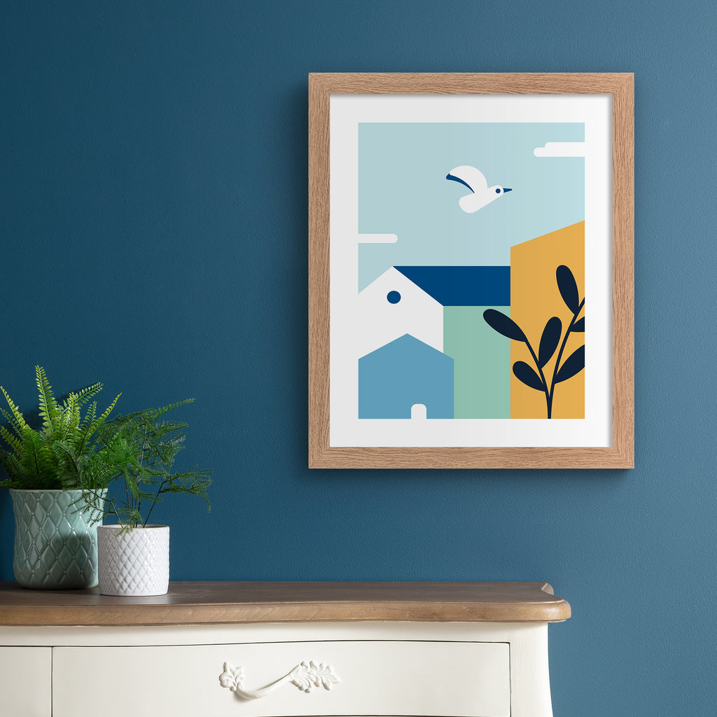 Minimalistic art print featuring a coastal bird flying over colourful houses. Art print is hung up on a dark blue wall.