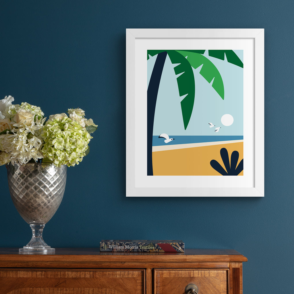 Minimalistic print featuring a palm tree standing in front of a beautiful coastal scene. Art print is hung up on a dark blue wall.