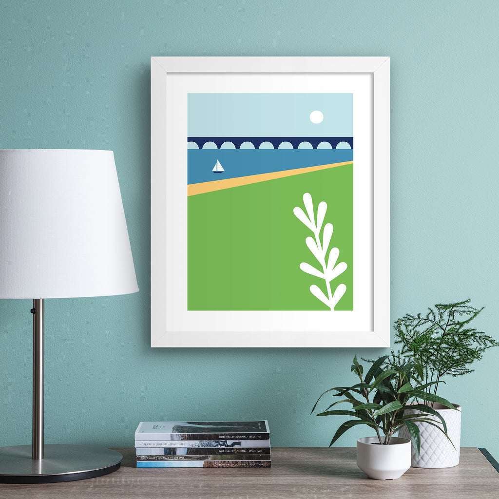 Minimalistic print containing a beautiful coastal scene, featuring a sun going down over a sailboat on still waters, near a bridge. Art print is hung up on a light blue wall.
