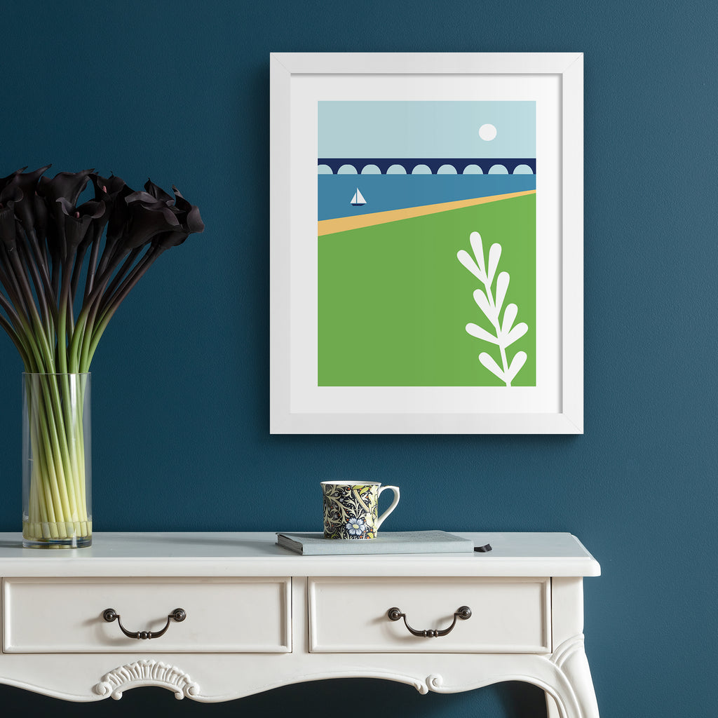 Minimalistic print containing a beautiful coastal scene, featuring a sun going down over a sailboat on still waters, near a bridge. Art print is hung up on a dark blue wall.